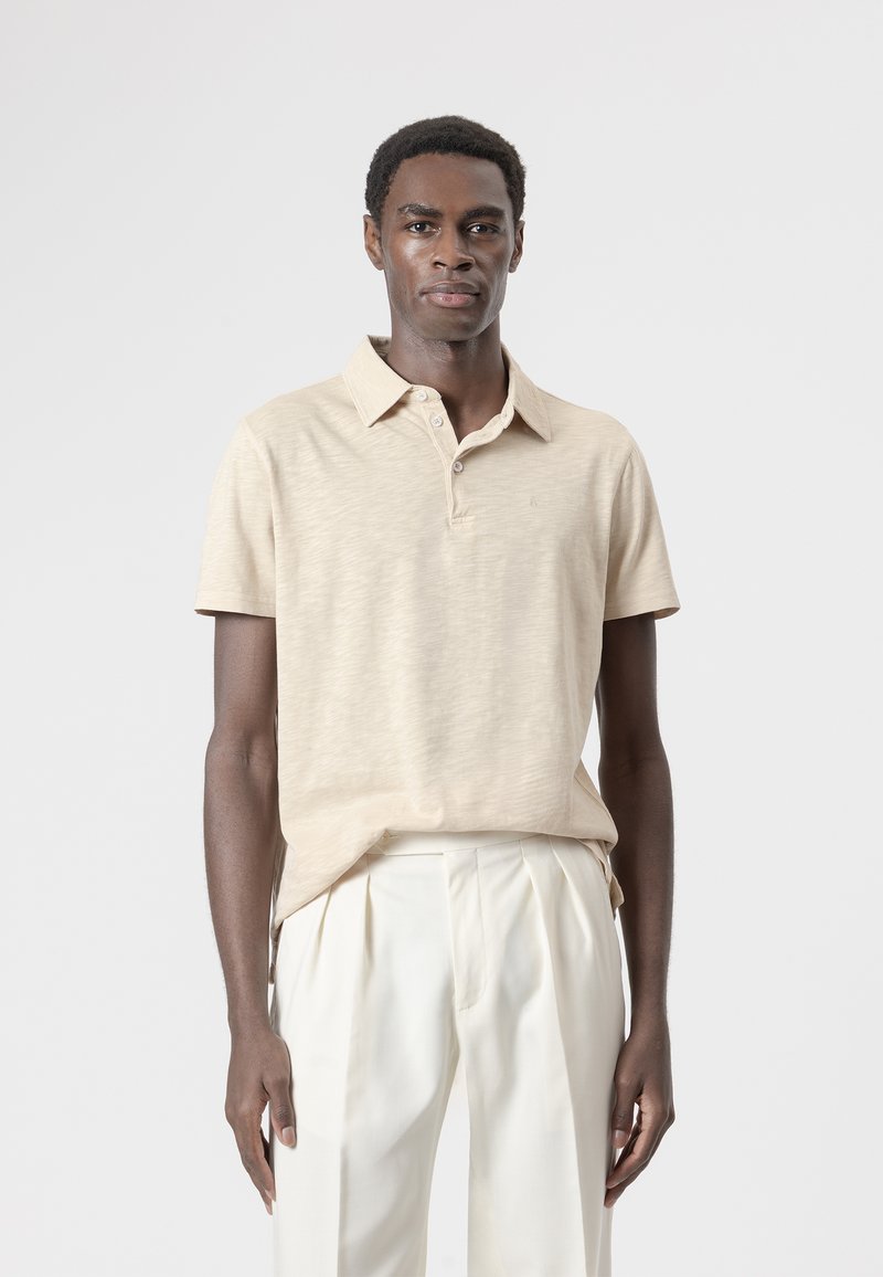 Hombre con una camisa tipo polo de manga corta beige metida en pantalones blancos plisados de talle alto, de pie sobre un fondo claro y liso.