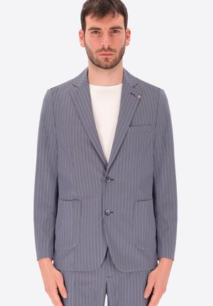 Uomo che indossa una giacca grigia testurizzata con due bottoni sopra una camicia bianca, in piedi davanti a uno sfondo grigio chiaro uniforme.