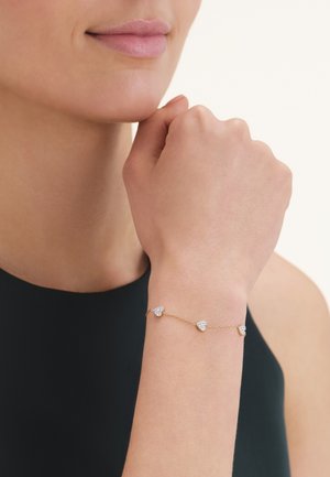 Persona che indossa un delicato braccialetto d'oro con tre piccoli charm a forma di cuore di diamante al polso contro uno sfondo chiaro.