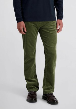Mann trägt olivgrüne Cordhose, dunkelbraune Lederloafers und ein marineblaues Langarmhemd und steht auf einem einfarbig hellen Hintergrund.