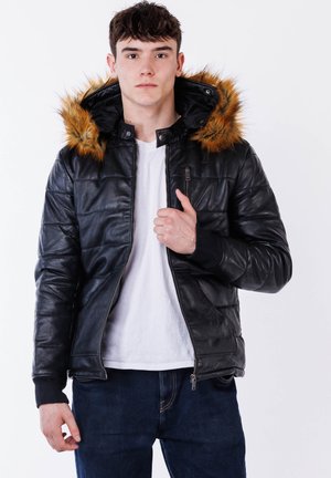 Młody mężczyzna ubrany w czarną skórzaną kurtkę z futrzanym kapturem, białą koszulkę i ciemne dżinsy, stojący na białym tle.