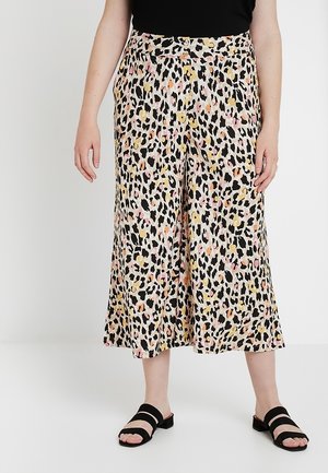 Vrouw draagt wijd uitlopende, cropped broek met meerkleurige luipaardprint en zwarte slip-on sandalen op een witte achtergrond.