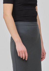 Jupe crayon gris asphalte avec une texture lisse, taille haute et une silhouette épurée. Haut noir à manches courtes partiellement visible.