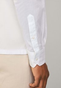 Biała koszula na długim rękawie z ostrym mankietem, z zapięciem na guziki i gładką fakturą. Koszula jest schowana w beżowe spodnie.