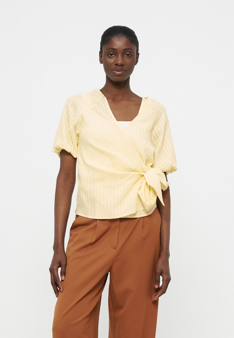 Γυναίκα που φοράει μπλούζα τύπου wrap σε απαλό κίτρινο με μανίκια φουσκωτά και καφέ παντελόνι με φαρδιά μπατζάκια, στέκεται μπροστά σε απλό λευκό φόντο.