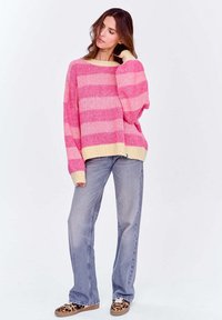 Oversized różowo-żółty sweter w paski z długim rękawem, w połączeniu z jasnoniebieskimi dżinsami i trampkami w panterkę.