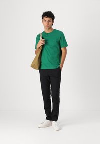 T-shirt en coton vert, à manches courtes, col rond, avec détail de logo. Associé à un pantalon noir et des baskets blanches. Sac beige porté à l'épaule.
