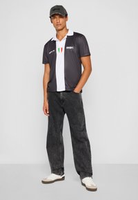Sort og hvid polo med korte ærmer og krave, der har et grønt, hvidt og rødt emblem. Sammen med mørk denim jeans og hvide sko.