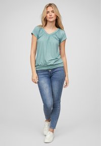 Lichtblauw kortmouwtop met een V-hals en geplooide details, gecombineerd met blauwe skinny jeans en witte sneakers. Glad textuur.