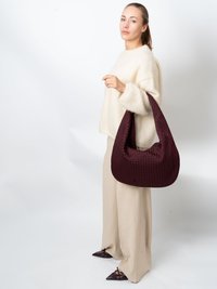 Burgundy flätad handväska med rundad form, texturerad yta och bred axelrem. Kombinerad med en kremfärgad tröja och beige vida byxor.