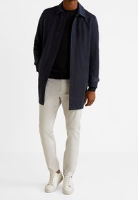Marineblauer leichter Mantel mit Knopfverschluss, langen Ärmeln, seitlichen Taschen, kombiniert mit grauen Hosen und weißen Turnschuhen. Glatte Stofftextur.