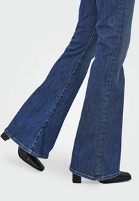 Jeans de mezclilla azul acampanados con un acabado oscuro y costuras visibles, combinados con zapatos de tacón bloque negro, destacando un diseño elegante y estructurado.