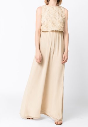 Femme portant une robe maxi beige sans manches avec un corsage floral texturé et une jupe fluide, présentée sur un fond blanc uni.