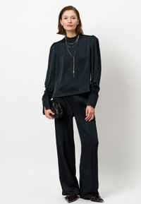Donkergroen blouse met een textuurpatroon, hoge halslijn en puffed mouwen, gecombineerd met losse zwarte broeken en een kleine zwarte clutch.