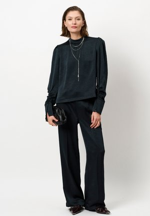 Donkergroen blouse met een textuurpatroon, hoge halslijn en puffed mouwen, gecombineerd met losse zwarte broeken en een kleine zwarte clutch.