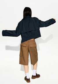 Lyhennetty tummansininen takki, jossa on leveät hihansuut ja elastinen helma, yhdistettynä ruskeisiin cargo-shortsihousuihin, joissa on taskut, valkoisiin sukkiin ja tummiin kenkiin.