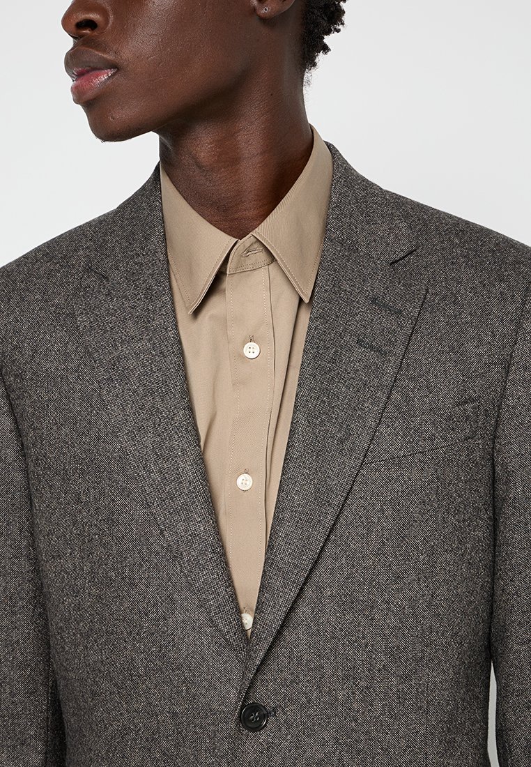 Mann in beiger Knopfleiste unter braunem strukturiertem Blazer, von Hals bis zur Mitte des Oberkörpers vor einem schlichten Hintergrund gezeigt.