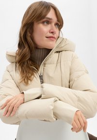 Beige puffer jacket met een capuchon, voorzien van een ritssluiting en geribbelde accenten. Soepele textuur en pluche isolatie zichtbaar in de mouwen.