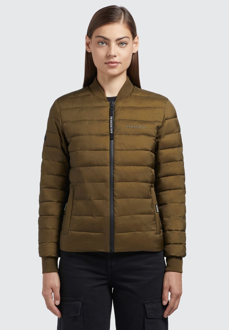 Chaqueta acolchada verde oliva con cierre de cremallera, puños acanalados y bolsillos laterales. Presenta un logo en el pecho y detalles de costura.