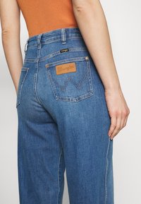 Modré džíny z denimu s rovný střih. Mají hnědou koženou nášivku s logem "Wrangler", měděné nýtky a dvojité zadní kapsy.