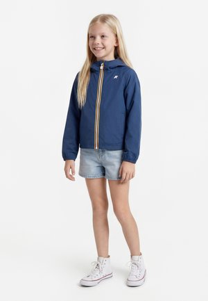 Pige stående iført en blå hættejakke, lyse denimbukser og hvide høje sneakers, smilende og kiggende let mod højre.