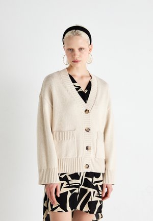 Beige knitted cardigan features a V-neck, button closure, two front pockets, and ribbed sleeve cuffs, worn over a black and white patterned dress.