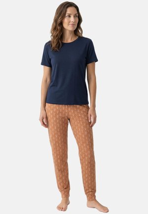 Kobieta stoi boso, ubrana w granatową koszulkę z krótkim rękawem i pomarańczowe spodnie w wzory na jednolitym jasnym tle.