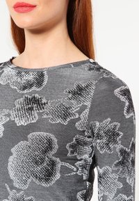 Woman with red hair wearing a gray long-sleeve top featuring large black and white floral patterns, shown from neck to mid-torso.