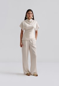 Krém színű szatén felső kámzsanyakkal és rövid ujjakkal, krém széles szárú nadrággal és arany színű papucs szandállal párosítva. Sima textúra és minimalista dizájn.