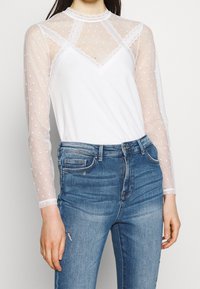 Top bianco con maniche lunghe in rete trasparente a pois e rifiniture in pizzo. Indossato con jeans in denim blu dal fit classico e leggera usura.