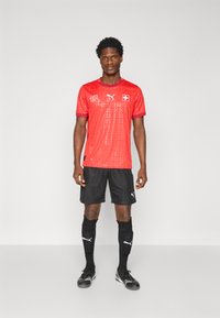 Rød fodboldtrøje med schweizisk flagdetalje, korte ærmer og mønstrede teksturer; parret med sorte shorts og sokker samt sorte fodboldstøvler.