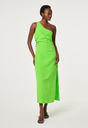 Frau trägt ein leuchtend grünes One-Shoulder-Midikleid mit seitlichen Knöpfen und einem Schlitz, kombiniert mit braunen Riemchensandalen, steht vor einem schlichten Hintergrund.