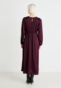 Pitkä violetti mekko, joka on valmistettu sileästä kankaasta. Siinä on puffihihat, kapea vyötärö ja avainaukko takana. Yhdistetty mustiin korkokenksiin.