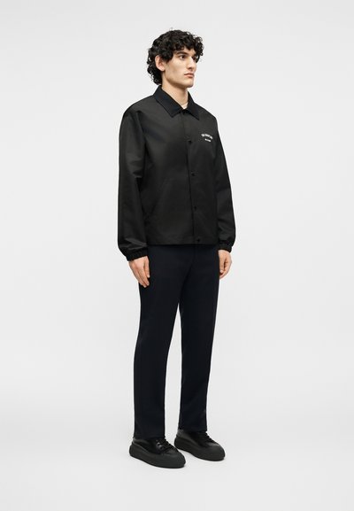 Νεαρός άνδρας με σγουρά μαλλιά στέκεται φορώντας μαύρο μπουφάν με κουμπιά, μαύρο παντελόνι και μαύρα αθλητικά παπούτσια σε ένα απλό λευκό φόντο.