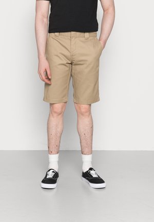 Homme portant un short beige mi-long, un t-shirt noir, des chaussettes blanches et des baskets noires, debout avec une main dans la poche devant un fond uni.