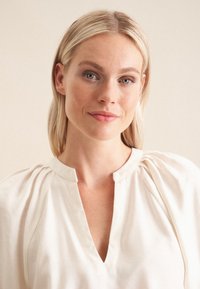 Hvid, linned bluse med V-udskæring og bløde plisseringer ved skuldrene. Glat tekstur og minimalistisk design.