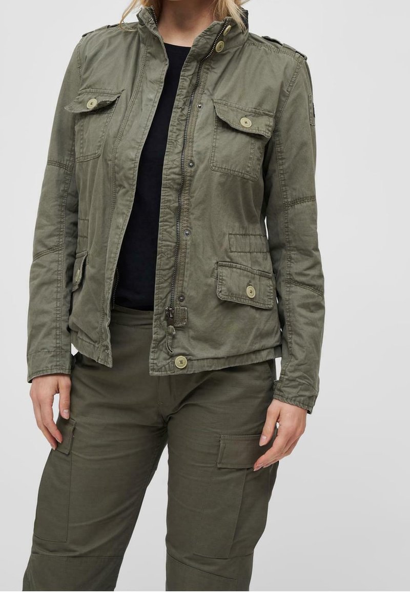 Personne portant une veste utilitaire vert olive avec des poches à rabat boutonnées et un pantalon cargo assorti, chemise noire en dessous, debout devant un fond uni.