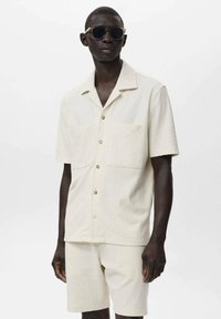 Kortærmet, beige skjorte med lodrette striber og to frontlommer. Sammen med matchende shorts. Modellen bærer mørke solbriller.