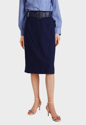 Donna che indossa una gonna a tubino blu navy fino al ginocchio, cintura nera con fibbia dorata testurizzata, camicia a righe blu e sandali con tacco alto color nudo.