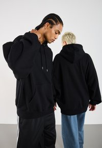 Sorte hættetrøjer i oversized pasform med frontlommer og snørehætter, parret med afslappede jeans, mod en ensfarvet baggrund.