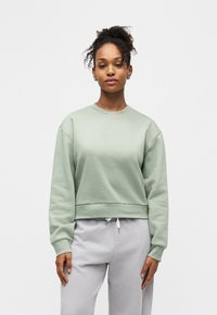 Femme aux cheveux bouclés attachés en chignon, portant un sweat-shirt vert sauge et un pantalon de survêtement gris clair, debout devant un fond clair uni.