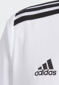 Camiseta atlética blanca con diseño de tres rayas negras en el hombro y un logo negro de Adidas en el frente. Textura de tela suave.