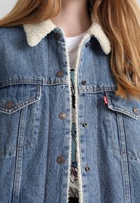 Denimjakke med krave foret med sherpa, lyseblå farve, knaplukning foran og to brystlommer. Rød logotag på venstre lomme.