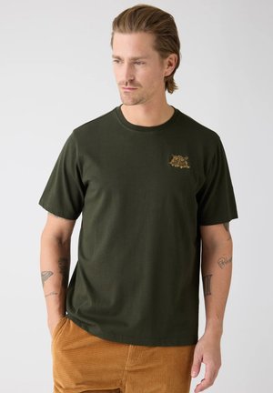 Uomo che indossa una maglietta verde scuro con un piccolo logo giallo a forma di gufo, pantaloni in velluto a coste color senape, in piedi con una mano in tasca, guarda a sinistra.