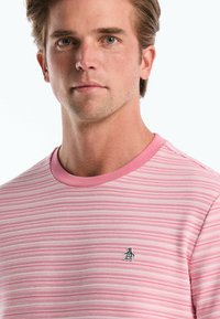 T-shirt à rayures roses avec un col rond et un petit logo noir sur la poitrine. Le tissu a une apparence texturée et une finition douce.