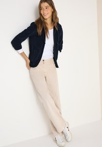 Femme appuyée contre un mur blanc, portant une veste zippée bleu marine, une chemise blanche, un pantalon beige et des baskets blanches avec des détails rouges.