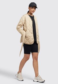 Beige gequiltte jas met een ontspannen pasvorm, voorzien van een drukknoopsluiting, gecombineerd met een zwart T-shirt en fietsbroek. Witte en beige sneakers.