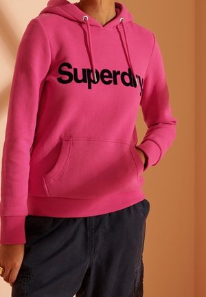 Osoba nosząca jasnoróżową bluzę z kapturem marki Superdry z przednią kieszenią i czarne spodnie typu cargo, stojąca na beżowym tle.