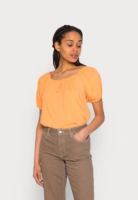 Orange puffærmet bluse med firkantet udskæring og samledetaljer; parret med brune højtaljede bukser med sidelommer.
