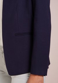 Gros plan sur une personne portant un blazer bleu marine foncé avec une poche passepoilée, un haut blanc et un pantalon clair, sur un fond neutre.